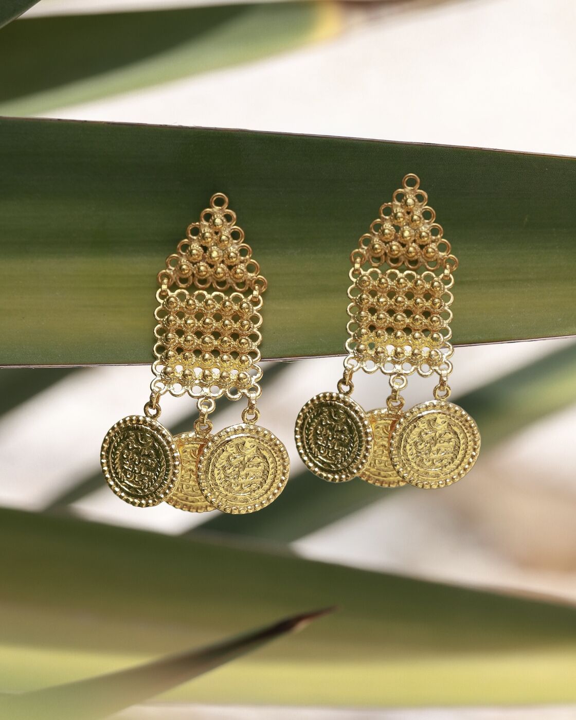 Gold coin drop earrings hanging against palm leaves
