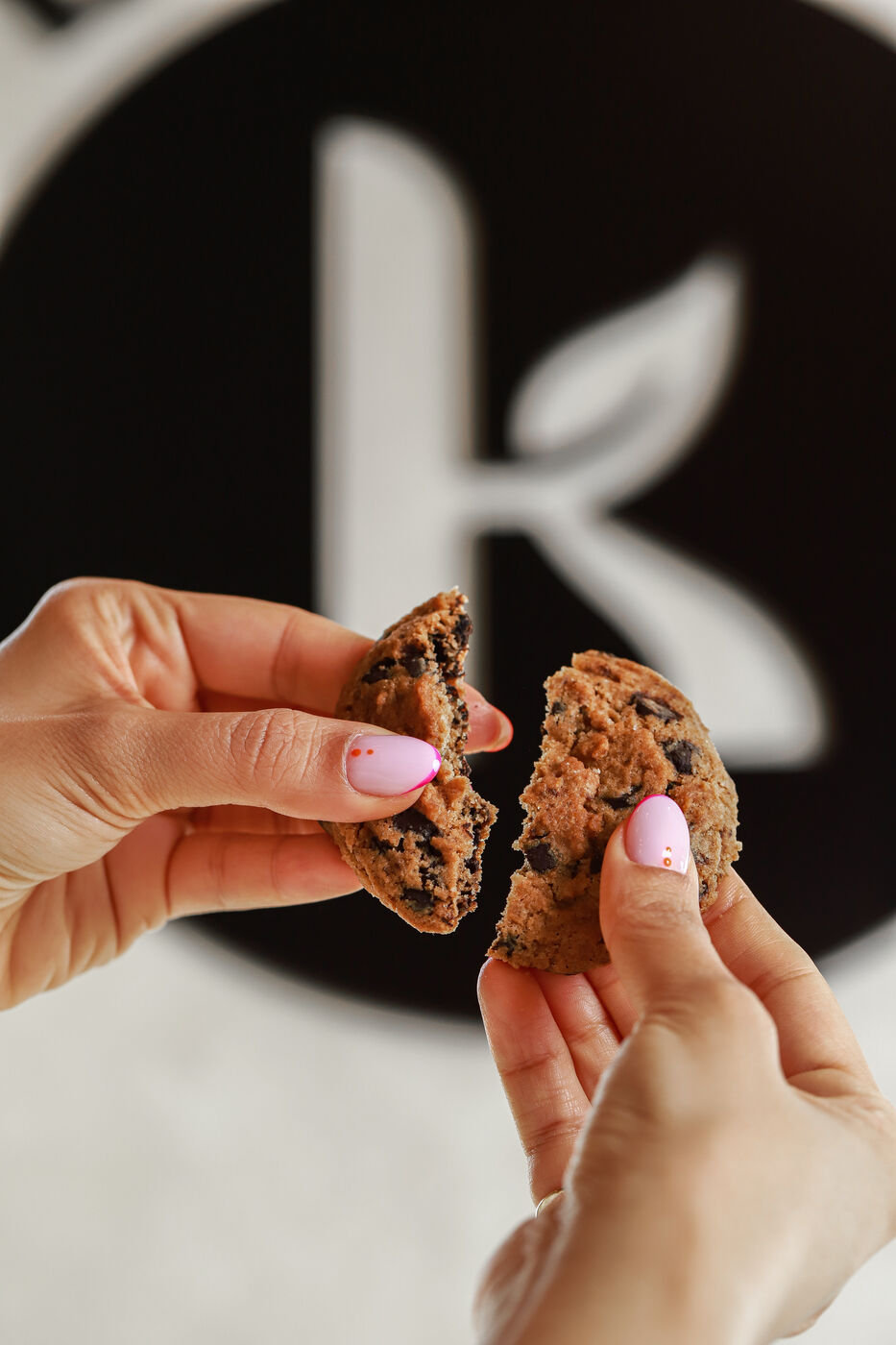 Hands breaking a chocolate chip cookie in front of a brand logo