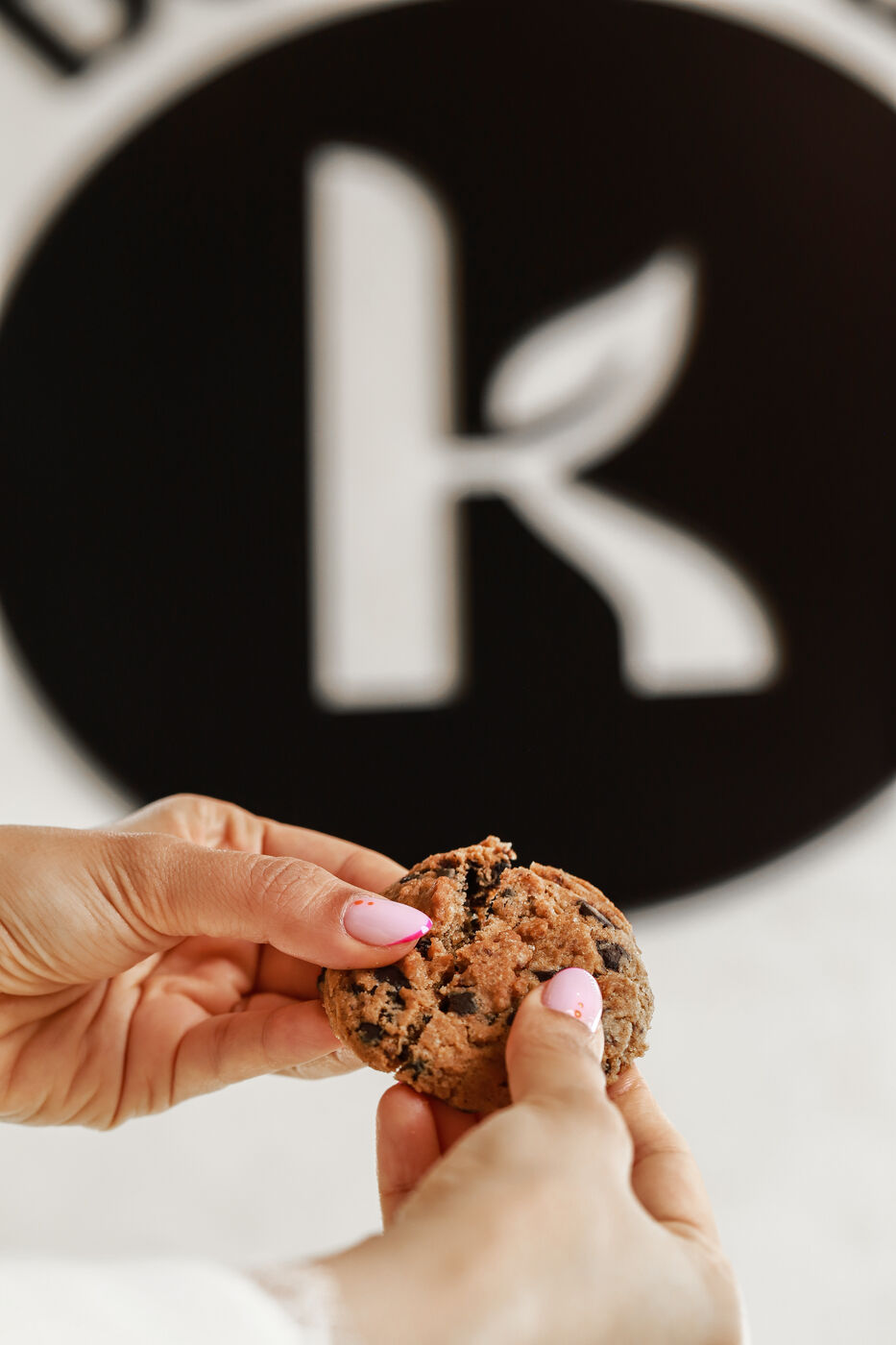 Close-up of hands holding a chocolate chip cookie in front of a brand logo