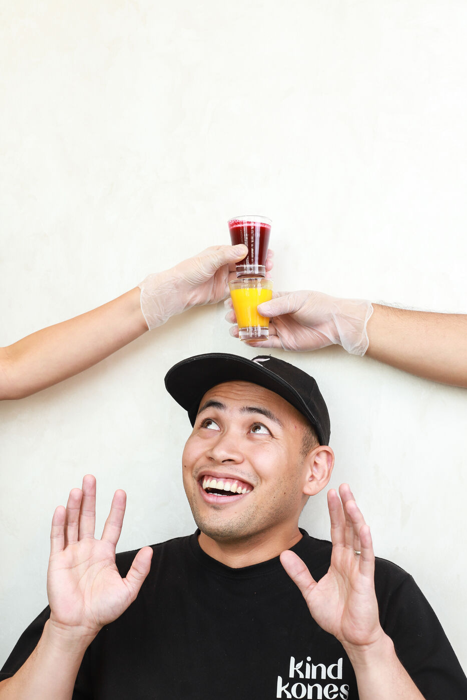 Man in a Kind Kones cap laughing as two shot glasses of juice are held above his head