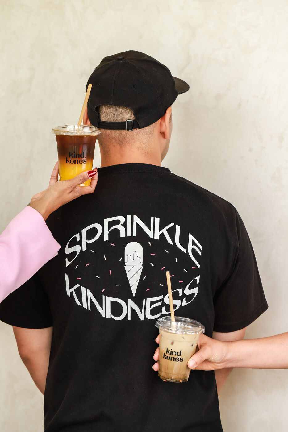 Back of a man's 'Sprinkle Kindness' t-shirt with two people holding Kind Kones iced drinks
